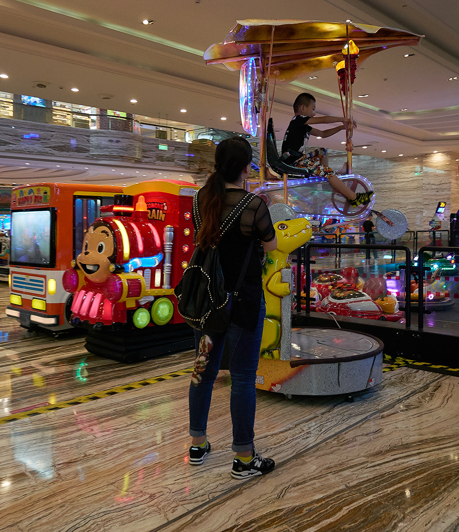 A kid riding a flying bicycle arcade game in the mall, while his parent watches